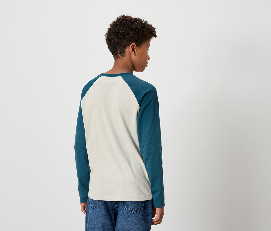 Ein junger Mann mit dem Rücken zur Kamera trägt ein zweifarbiges Langarmshirt und Jeans.