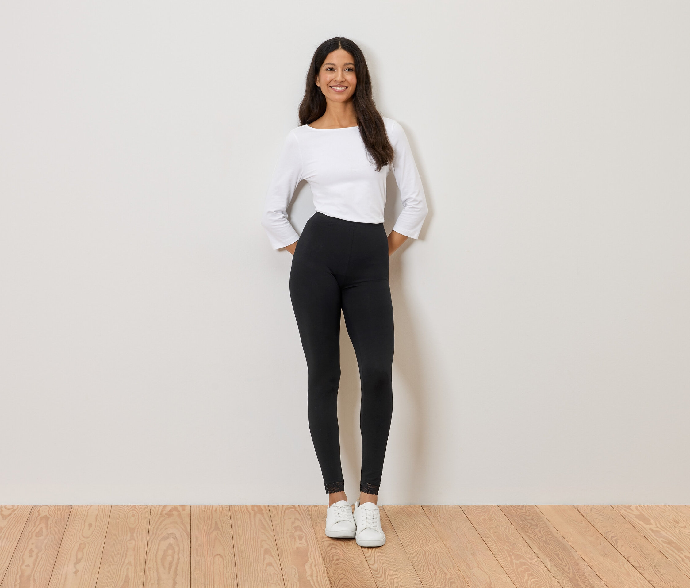 Frau steht vor einer Wand, trägt ein weißes Oberteil, schwarze Leggings und weiße Turnschuhe.
