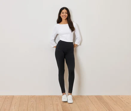 Frau steht vor einer Wand, trägt ein weißes Oberteil, schwarze Leggings und weiße Turnschuhe.