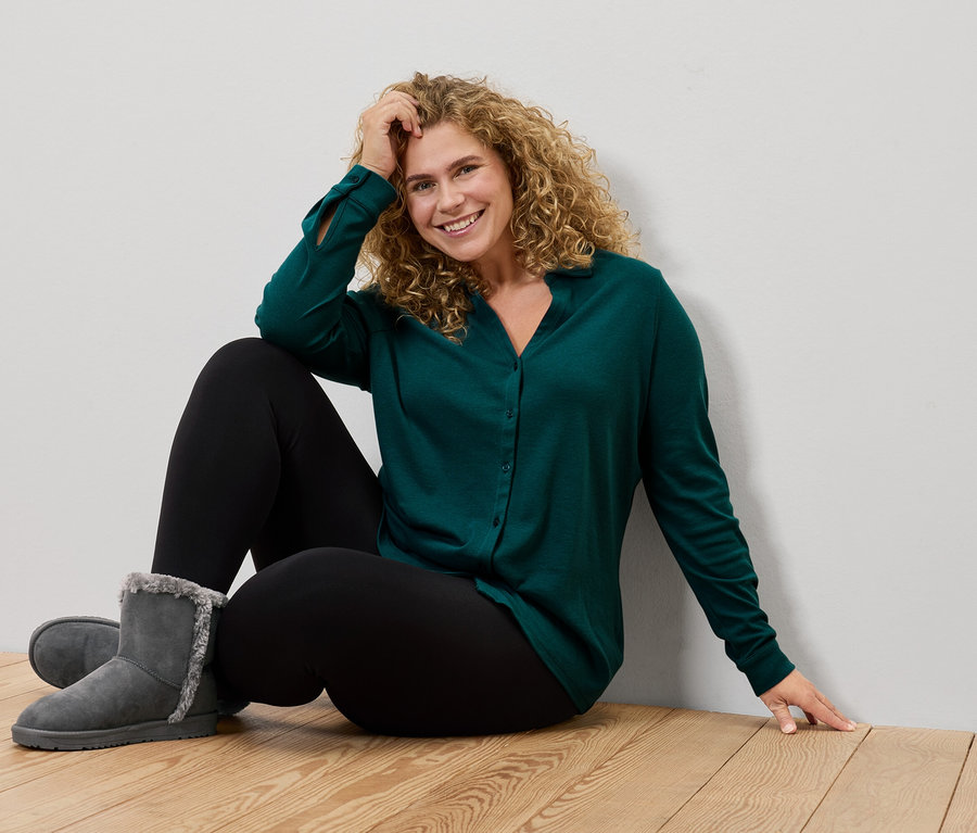 Frau sitzt auf dem Boden, trägt ein grünes Hemd, schwarze Leggings und graue Stiefel.