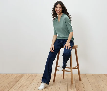 Frau sitzt auf einem Hocker und trägt ein Feinstrick-Polo, Jeans und Turnschuhe.