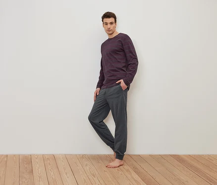 Ein Mann steht barfuß auf einem Holzboden und trägt ein lila gestreiftes Langarmshirt und eine graue Jogginghose.
