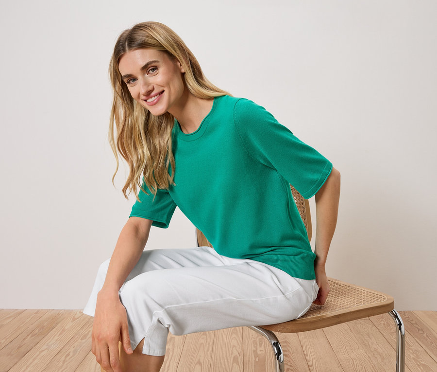 Frau sitzt auf einem Stuhl und trägt ein grünes Kurzarm-Shirt und eine weiße Hose.