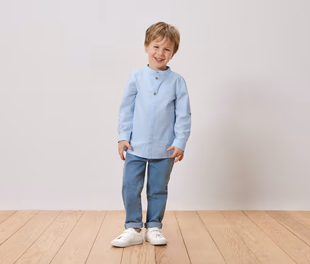 Ein lächelnder Junge mit blonden Haaren steht vor einer weißen Wand. Er trägt ein hellblaues Hemd, Jeans und weiße Schuhe.