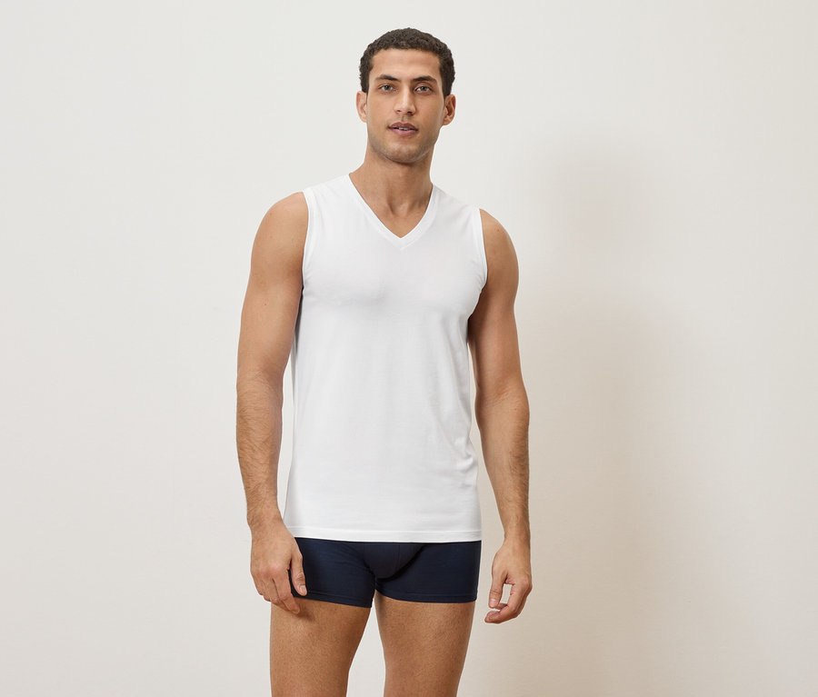 Mann steht in Unterwäsche und weißem Tanktop vor einer schlichten Wand.