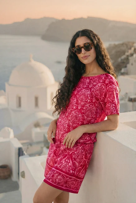 Frau mit Sonnenbrille, Blusenshirt mit Alloverprint und bedruckten Webshorts, lehnt an einer Wand. Sie trägt Creolen und einen transparenten Ring.