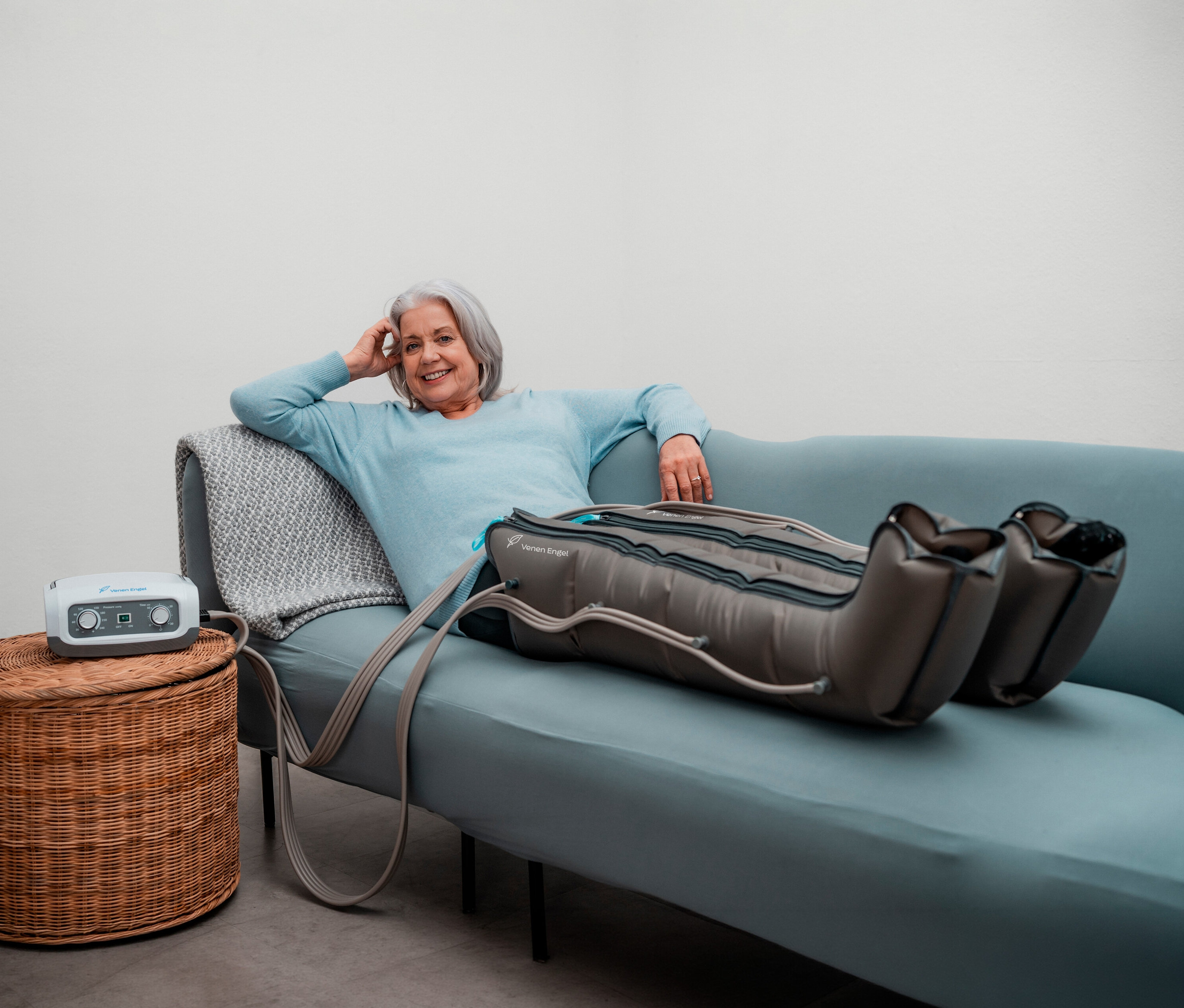 Frau entspannt mit Venen Engel 4 - Basic Gleitwellenmassage-Gerät mit Beinmanschette auf Sofa. Gerät steht auf Korbhocker.