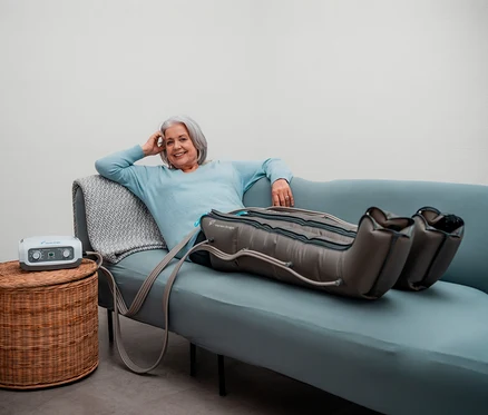 Frau entspannt mit Venen Engel 4 - Basic Gleitwellenmassage-Gerät mit Beinmanschette auf Sofa. Gerät steht auf Korbhocker.