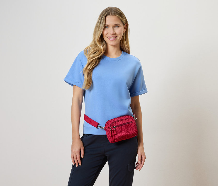 Frau trägt ein hellblaues T-Shirt und eine rote Bauchtasche vor einem neutralen Hintergrund.
