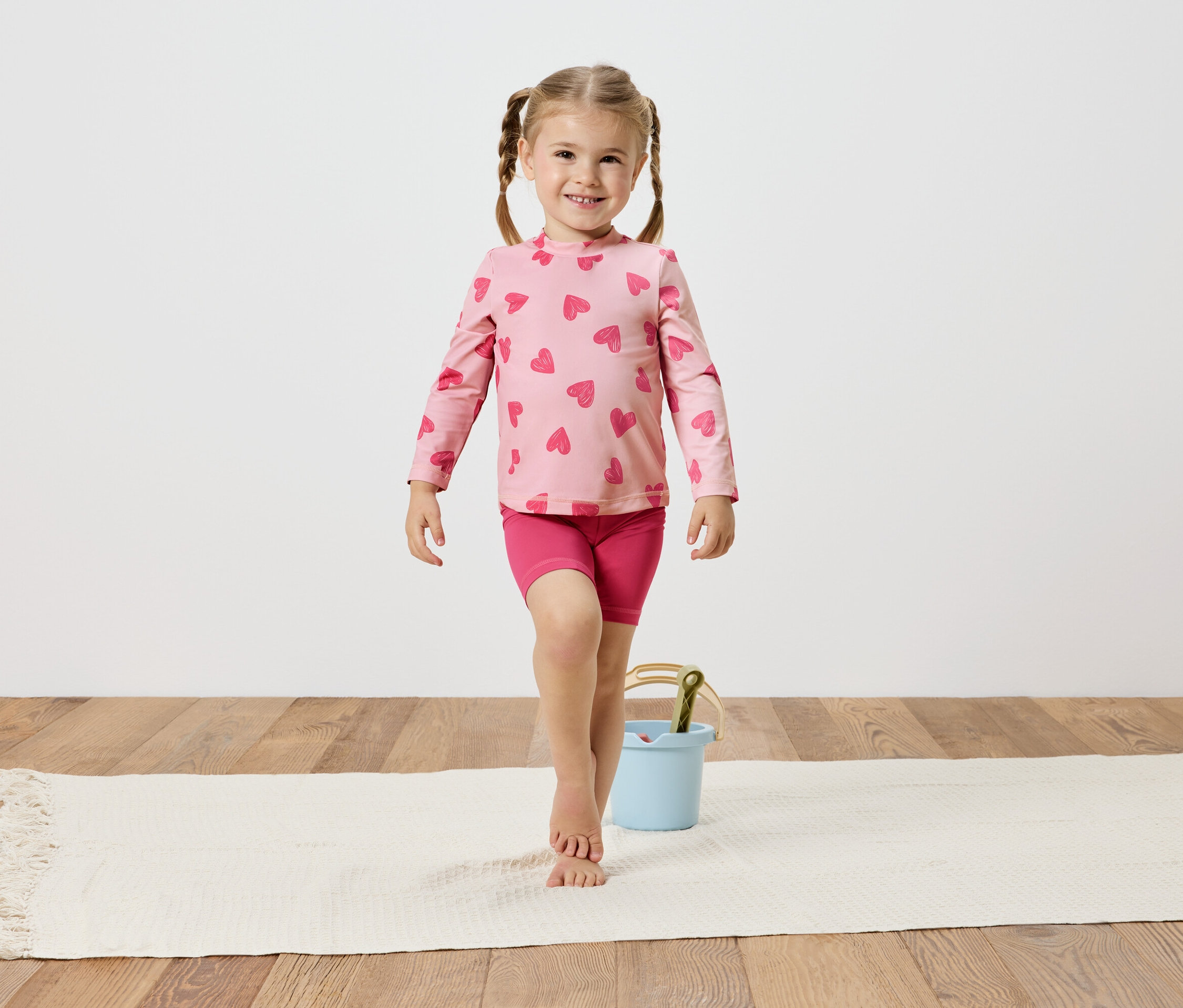 Mädchen mit Zöpfen, trägt ein rosa Herzchenoberteil und Shorts, balanciert auf einem weißen Teppich neben einem hellblauen Eimer.