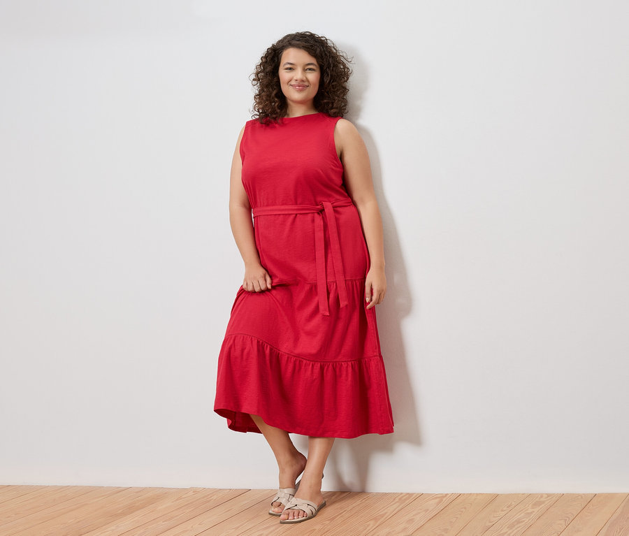 Eine Frau mit lockigem Haar steht vor einer weißen Wand und trägt ein rotes, ärmelloses Kleid mit Gürtel und Sandalen.