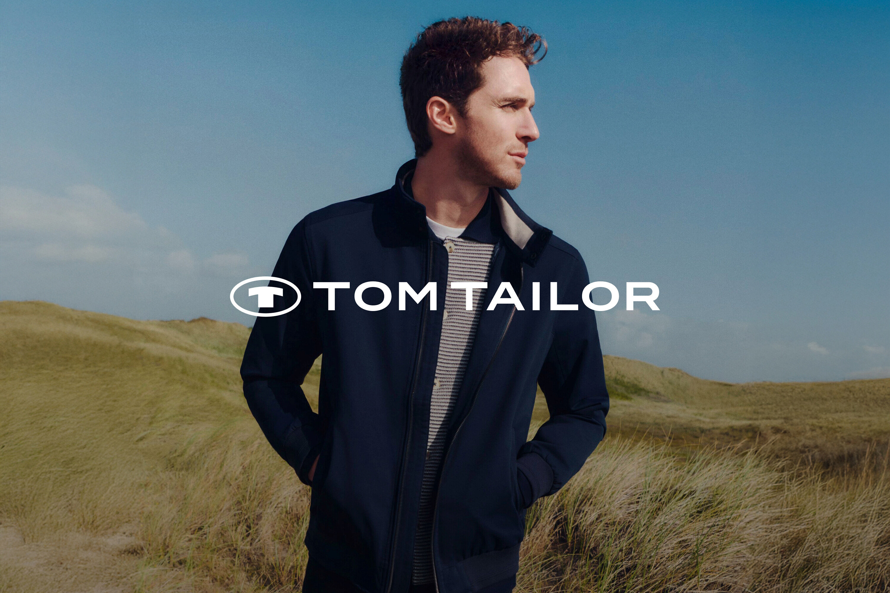 Mann mit dunkelblauer Jacke und gestreiftem Hemd vor Graslandschaft und blauem Himmel.