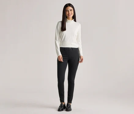 Frau mit langem, dunklem Haar trägt einen weißen Rollkragenpullover, eine schwarze Hose und schwarze Schuhe.