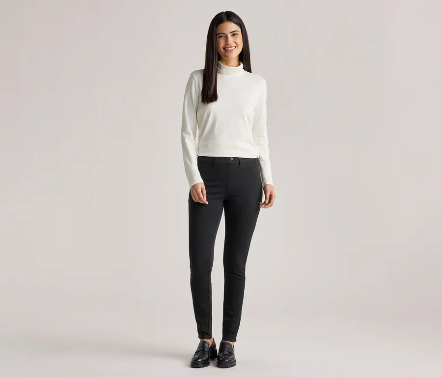 Frau mit langem, dunklem Haar trägt einen weißen Rollkragenpullover, eine schwarze Hose und schwarze Schuhe.