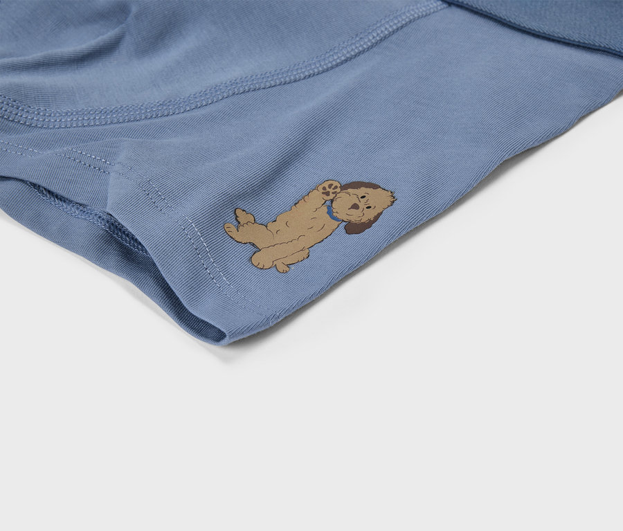 Nahaufnahme eines blauen Stoffes, möglicherweise Unterwäsche, mit einer hellbraunen Cartoon-Hund-Illustration, die auf dem Rücken liegt und die Pfoten hochhält. Der Hund hat dunkelbraune Ohren und ein blaues Halsband.