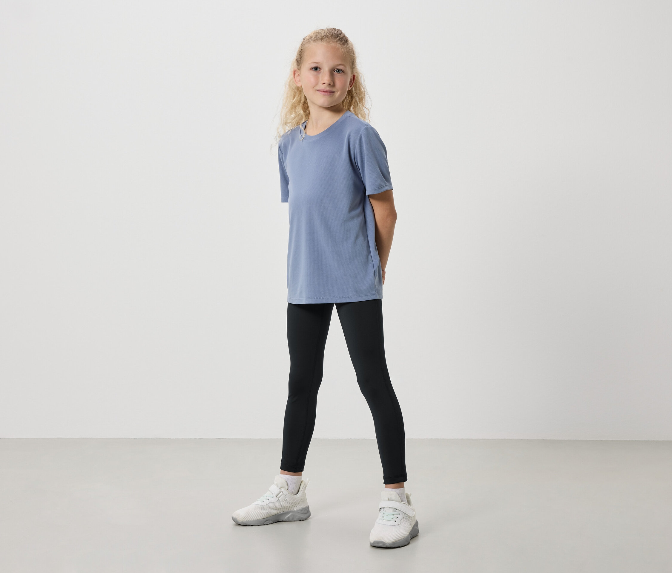 Mädchen steht frontal in blauem T-Shirt, schwarzen Leggings und weißen Turnschuhen vor einem hellen Hintergrund.