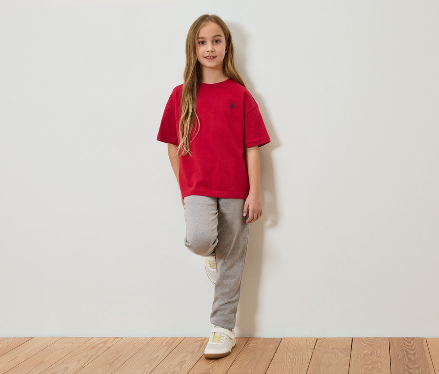 Mädchen mit langem braunem Haar, rotem T-Shirt und grauer Jogginghose, das an einer weißen Wand lehnt.