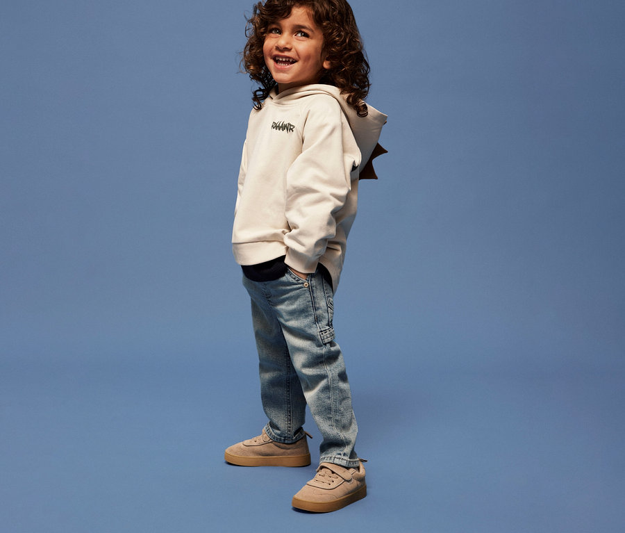 Junge mit lockigem Haar trägt ein NAME IT Kinder-Sweatshirt mit 3D-Dino-Applikation, Jeans und Turnschuhe vor blauem Hintergrund.