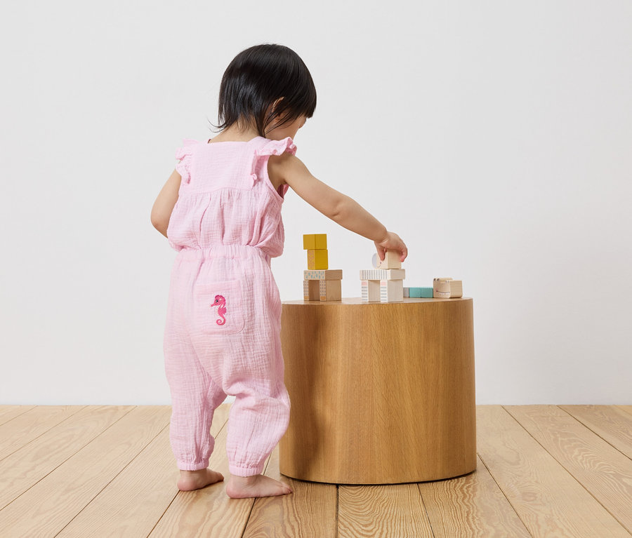 Kleines Kind in einem rosa Overall mit Seepferdchen-Motiv spielt mit Bauklötzen auf einem runden Holztisch.