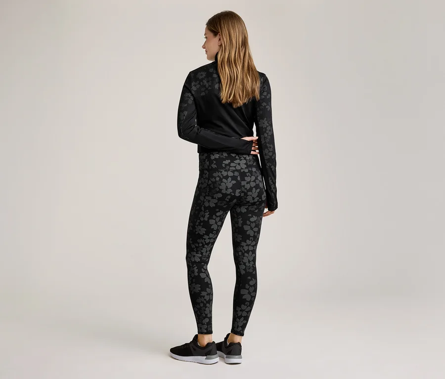 Rückansicht einer Frau mit langen, blonden Haaren in schwarzer Sportbekleidung mit Blumenmuster, bestehend aus Leggings und Oberteil.
