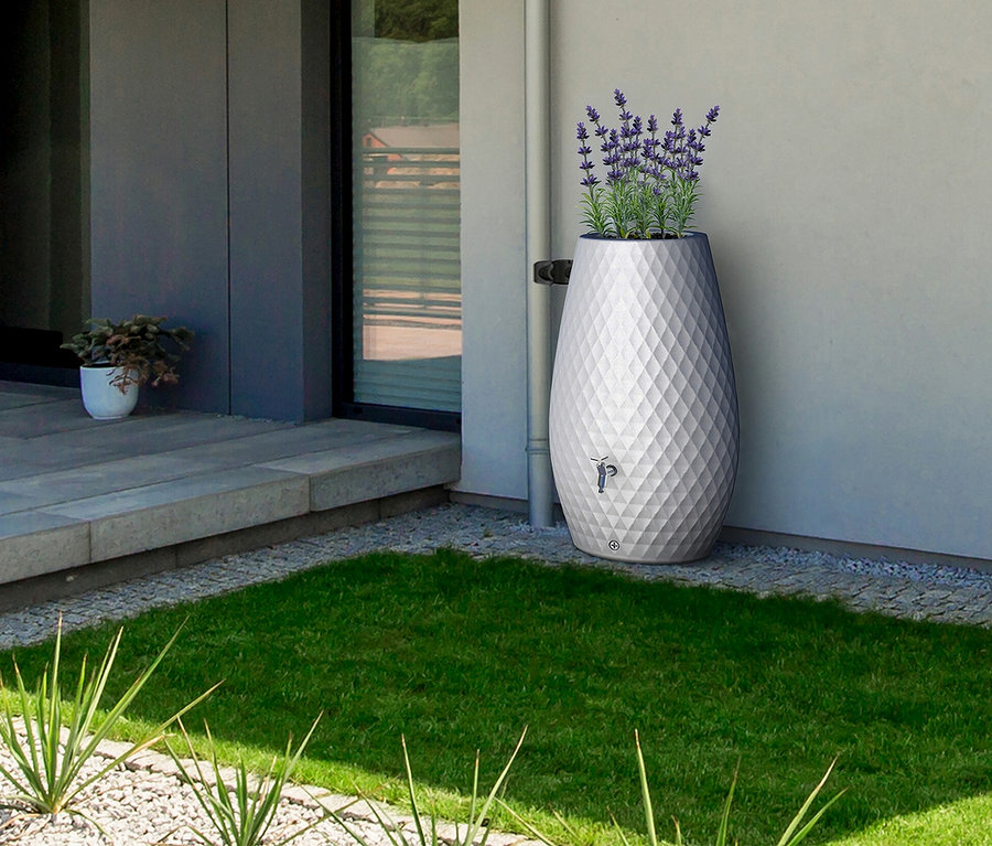 Eine große, weiße Regentonne mit Rautenmuster und lila Blumen steht auf Kies neben einem Rasen vor einem Haus.