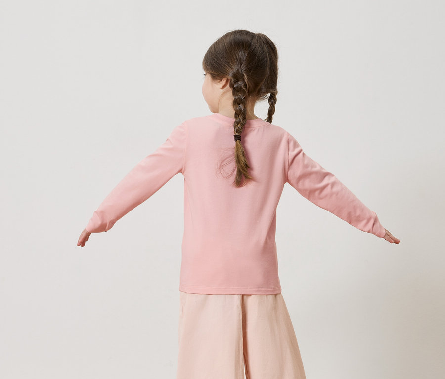 Rückenansicht eines kleinen Mädchens mit Zöpfen, das ein rosa Langarmshirt und eine rosafarbene Hose trägt, die Arme sind seitlich ausgestreckt.