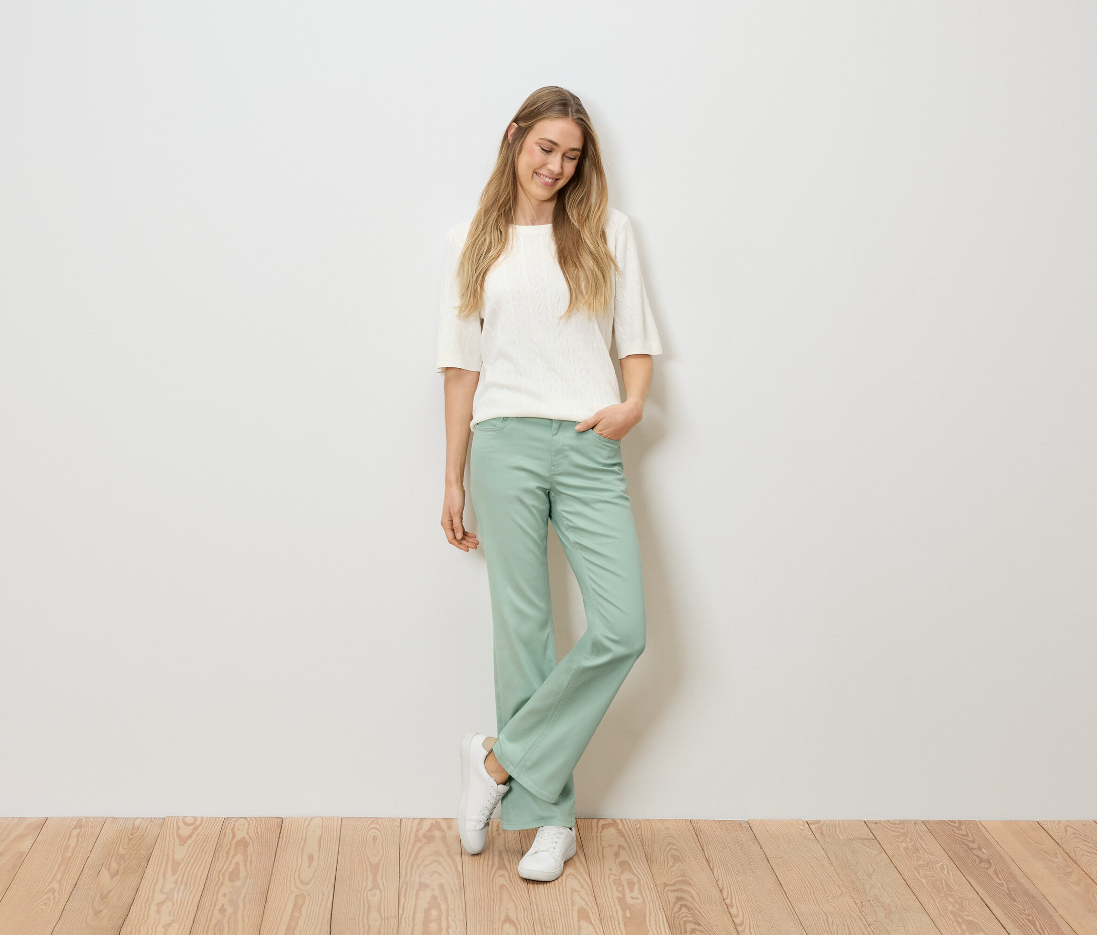 Frau mit blonden Haaren, die ein weißes Oberteil, eine grüne Hose und weiße Turnschuhe trägt, steht vor einer weißen Wand auf einem Holzboden.