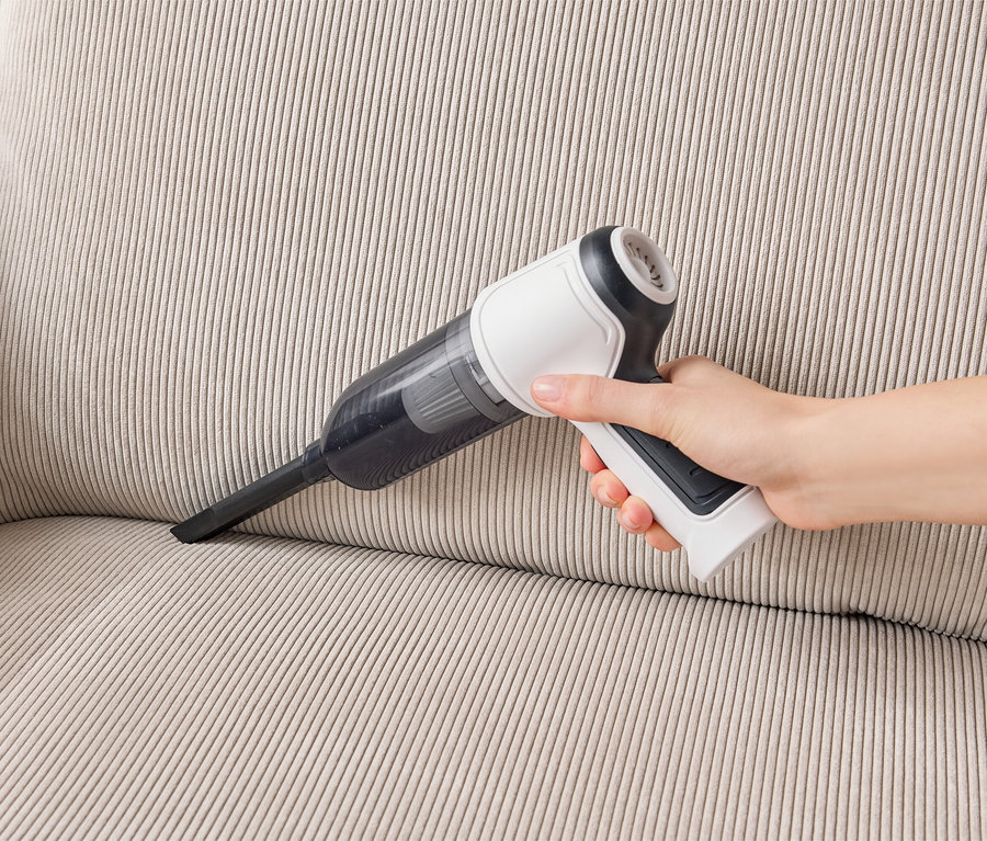 Eine Hand hält einen weißen und schwarzen Handstaubsauger und saugt eine beigefarbene Couch ab.