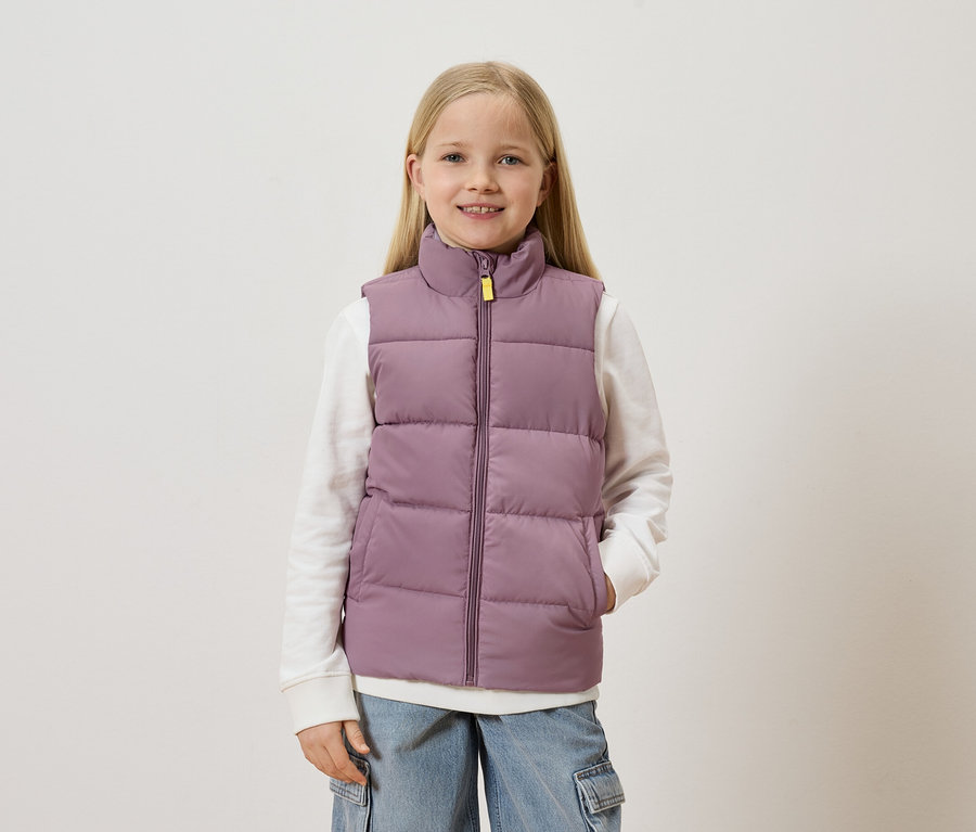 Mädchen mit blonden Haaren trägt eine lila Weste über einem weißen Pullover und blauen Jeans mit Taschen.