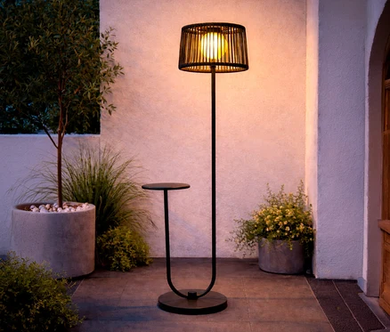Eine anthrazitfarbene Leco Solar Stehleuchte mit integriertem Tisch steht auf einer Terrasse neben Pflanzen.