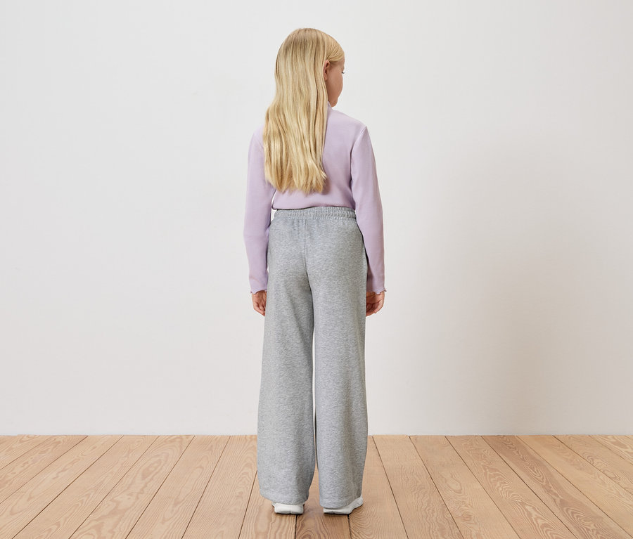 Blick von hinten auf ein blondes Mädchen mit helllila Pullover und grauer, weiter Hose vor einer weißen Wand.