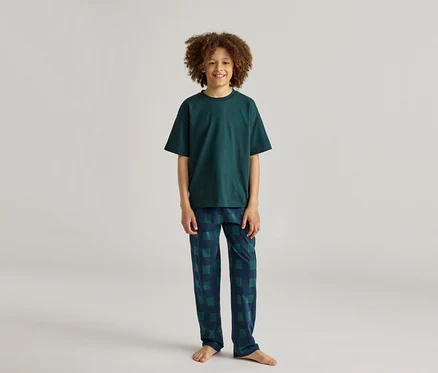 Ein lächelnder Junge mit lockigem Haar trägt einen dunkelgrünen Pyjama mit karierter Hose.