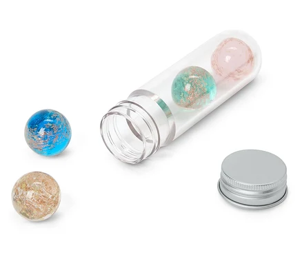 Vier glitzernde Kugeln in Blau, Türkis, Rosa und Beige, zwei davon in einer offenen, durchsichtigen Plastikröhre und zwei daneben. Ein silberner Deckel liegt neben der Röhre.
