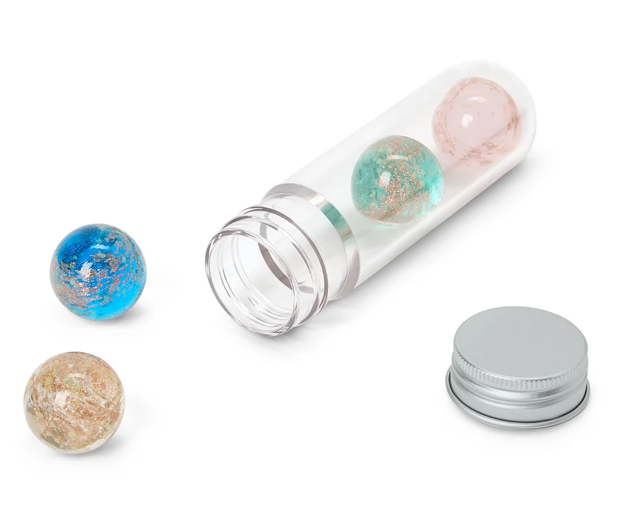 Vier glitzernde Kugeln in Blau, Türkis, Rosa und Beige, zwei davon in einer offenen, durchsichtigen Plastikröhre und zwei daneben. Ein silberner Deckel liegt neben der Röhre.