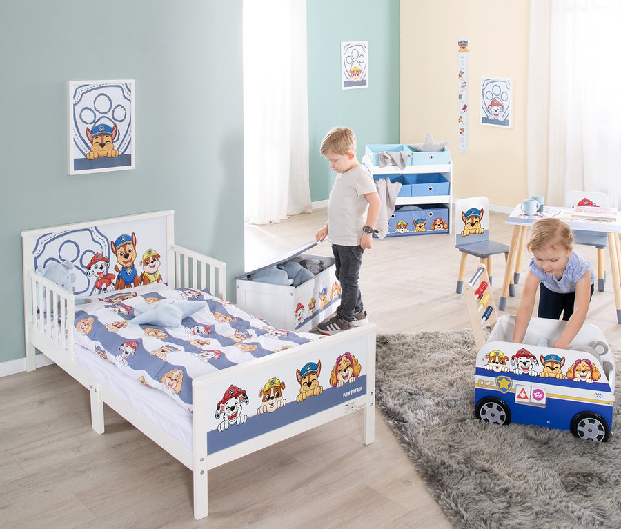 Kinderzimmer mit Paw Patrol-Motiven: Junge steht auf Spielzeugkiste neben Bett, Mädchen spielt mit Spielzeugbus auf Teppich.