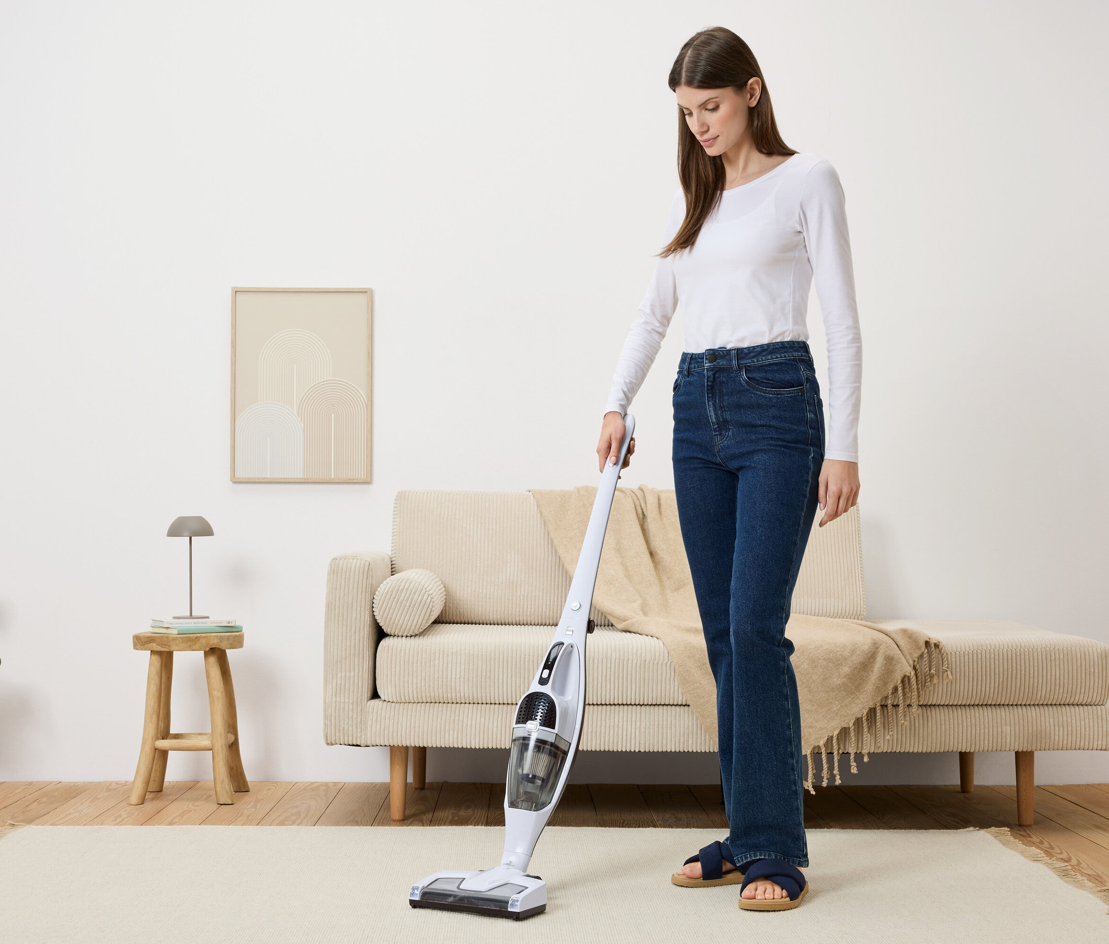 Frau staubsaugt mit einem weißen Akku-Staubsauger einen beigen Teppich in einem Wohnzimmer.
