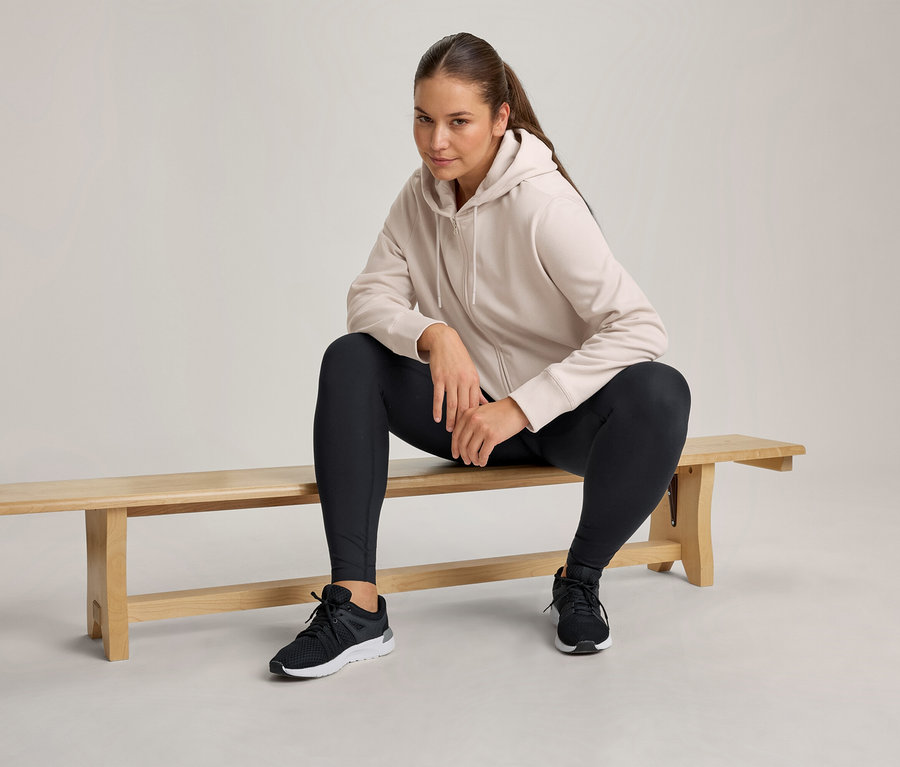 Frau sitzt auf einer Holzbank, trägt einen hellbeigen Kapuzenpullover mit Reißverschluss, schwarze Leggings und schwarze Sportschuhe.