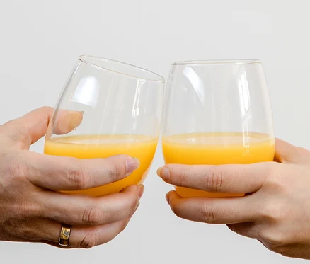 Zwei Hände stoßen mit Mäser Wassergläsern »Il Premio« an, die mit Orangensaft gefüllt sind. Eine Hand trägt einen Ring.