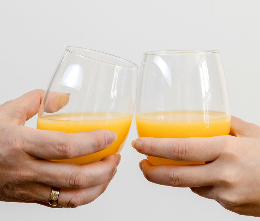 Zwei Hände stoßen mit Mäser Wassergläsern »Il Premio« an, die mit Orangensaft gefüllt sind. Eine Hand trägt einen Ring.