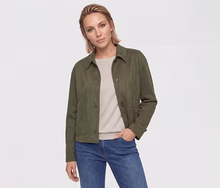 Eine Frau mit kurzen, hellbraunen Haaren trägt eine dunkelgrüne Jacke, ein beiges Oberteil und blaue Jeans. Ihre Hand steckt in der Hosentasche.