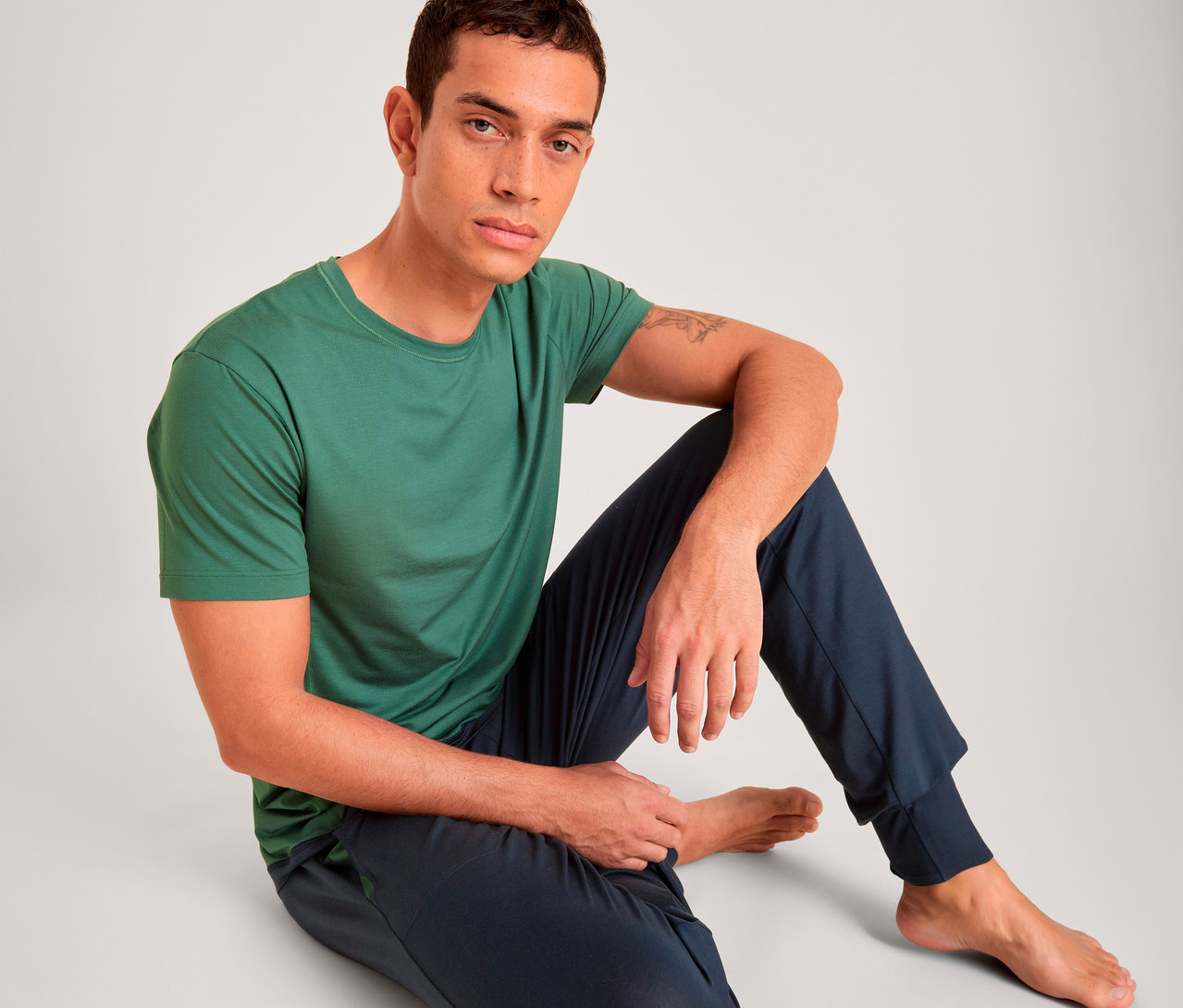 Mann sitzt auf dem Boden, trägt ein grünes T-Shirt und dunkelblaue Jogginghose, blickt in die Kamera.