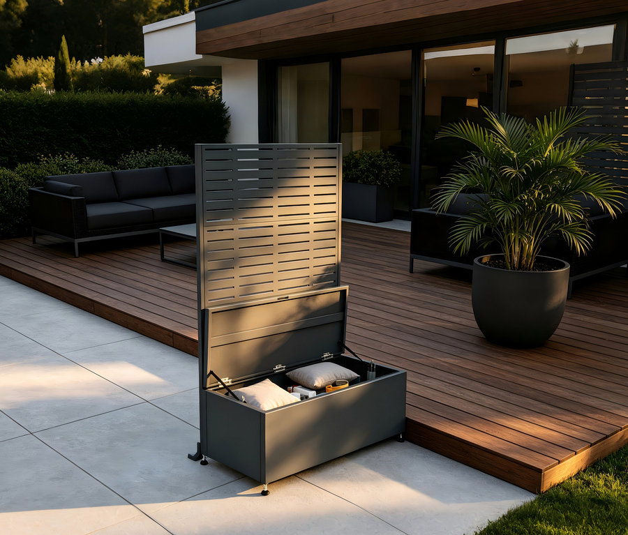 Dunkelgraue Outdoor-Aufbewahrungsbox mit offenem Deckel und integriertem Lamellen-Sichtschutz auf einer Terrasse. Kissen sind in der Box. Dahinter befindet sich eine Holzterrasse mit Sofa und Topfpflanze.
