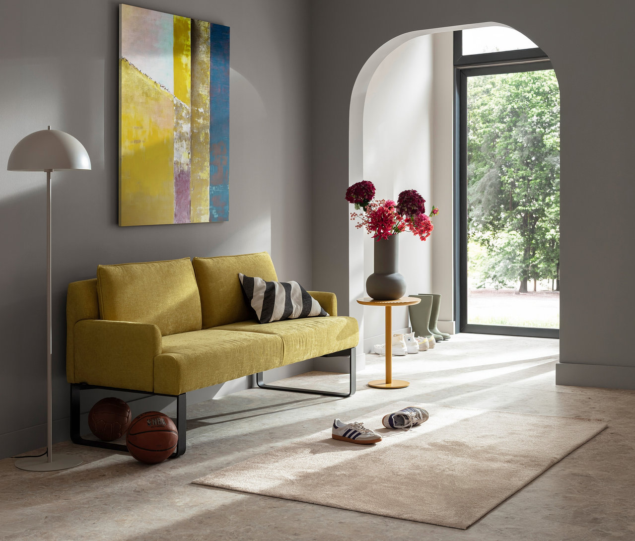 Gelbes Zweisitzer-Sofa mit einer schwarz-weiß gestreiften SCHÖNER WOHNEN - Kollektion Zierkissenhülle »JUNCTION« vor grauer Wand mit Bild. Bogen führt in Garten.