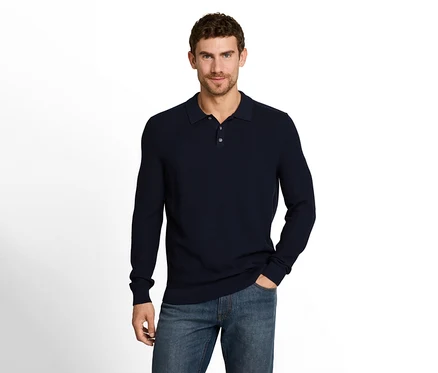Mann steht, trägt ein dunkelblaues Langarm-Poloshirt und blaue Jeans, eine Hand in der Tasche, blickt in die Kamera.
