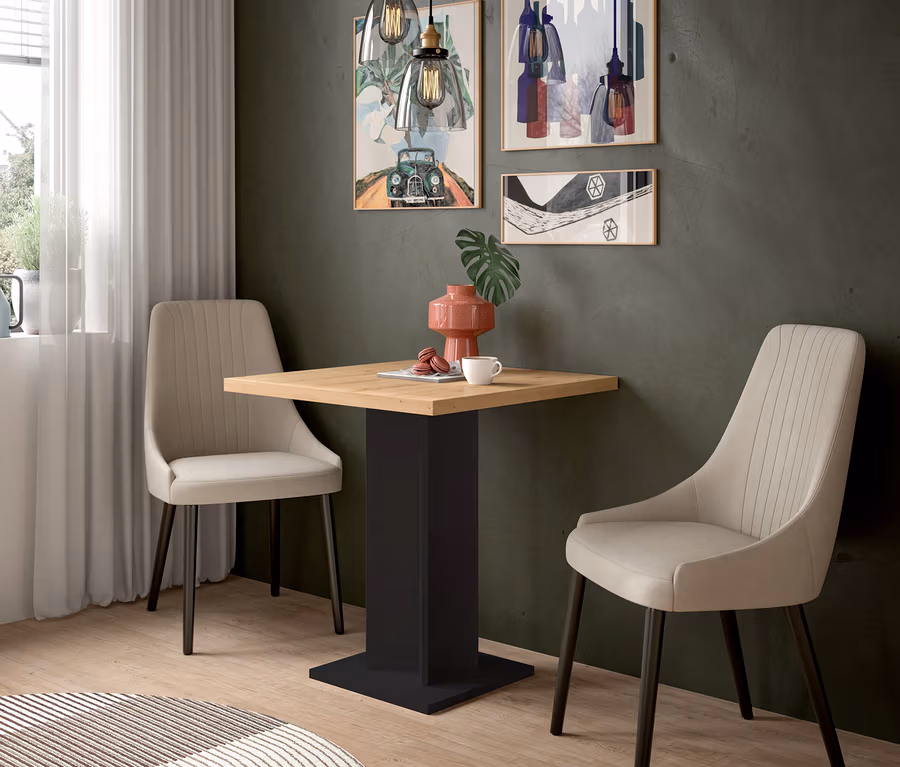 Ein Essbereich mit einem quadratischen Holztisch mit schwarzem Sockel und zwei beigen Stühlen. Auf dem Tisch stehen eine rosa Vase, eine Tasse und Macarons. An der dunkelgrünen Wand hängen drei gerahmte Bilder, darüber zwei Pendelleuchten. Links ist ein Fenster mit weißen Vorhängen zu sehen, am Boden liegt ein runder Teppich.