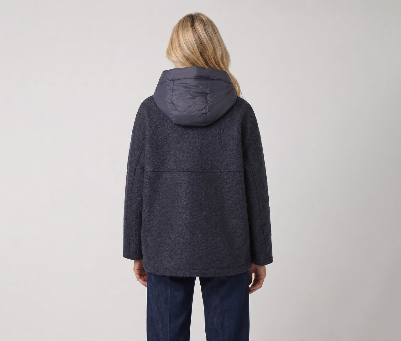 Rückenansicht einer Person mit blonden Haaren, die eine dunkelblaue, strukturierte Jacke mit gepolsterter Kapuze und dunkelblaue Jeans trägt.