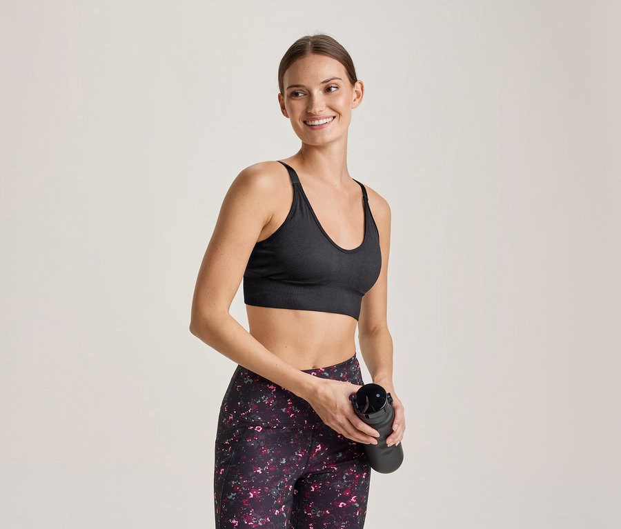 Lächelnde Frau in schwarzem Sport-BH und gemusterten Leggings hält eine schwarze Trinkflasche.
