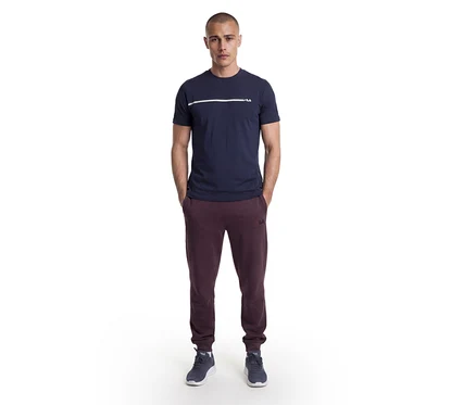 Mann steht mit Händen in den Taschen, trägt ein dunkelblaues T-Shirt mit weißem Streifen und Fila-Logo, bordeauxrote Fila-Jogginghose und dunkelblaue Sneaker.
