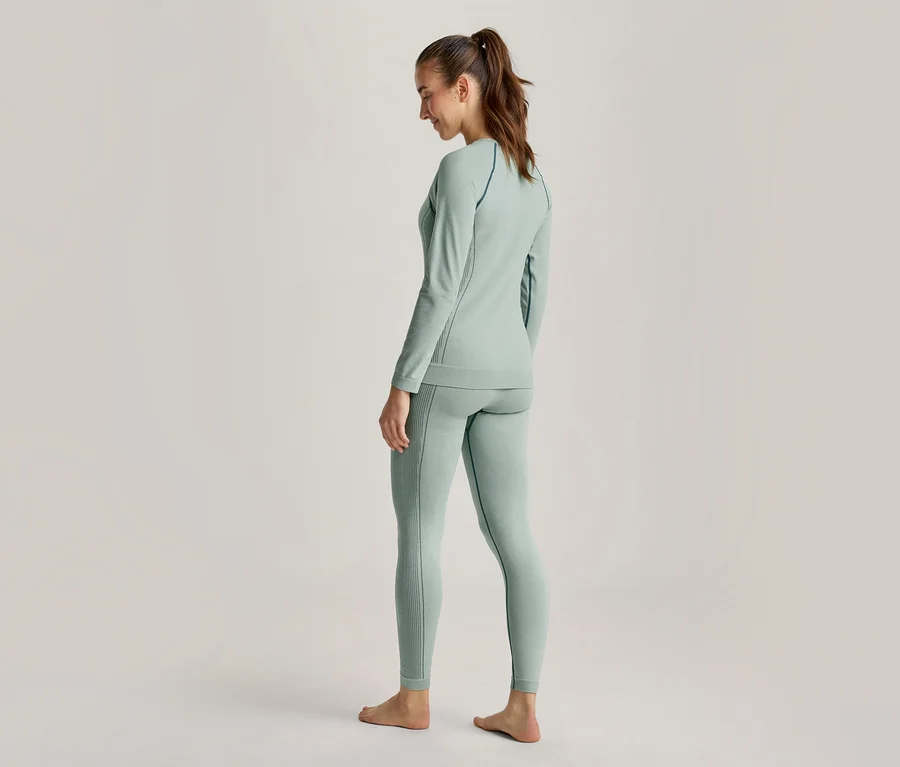 Frau von hinten, leicht zur Seite gedreht, trägt ein hellgrünes Langarmshirt und passende Leggings, die Haare zum Pferdeschwanz gebunden, barfuß stehend.