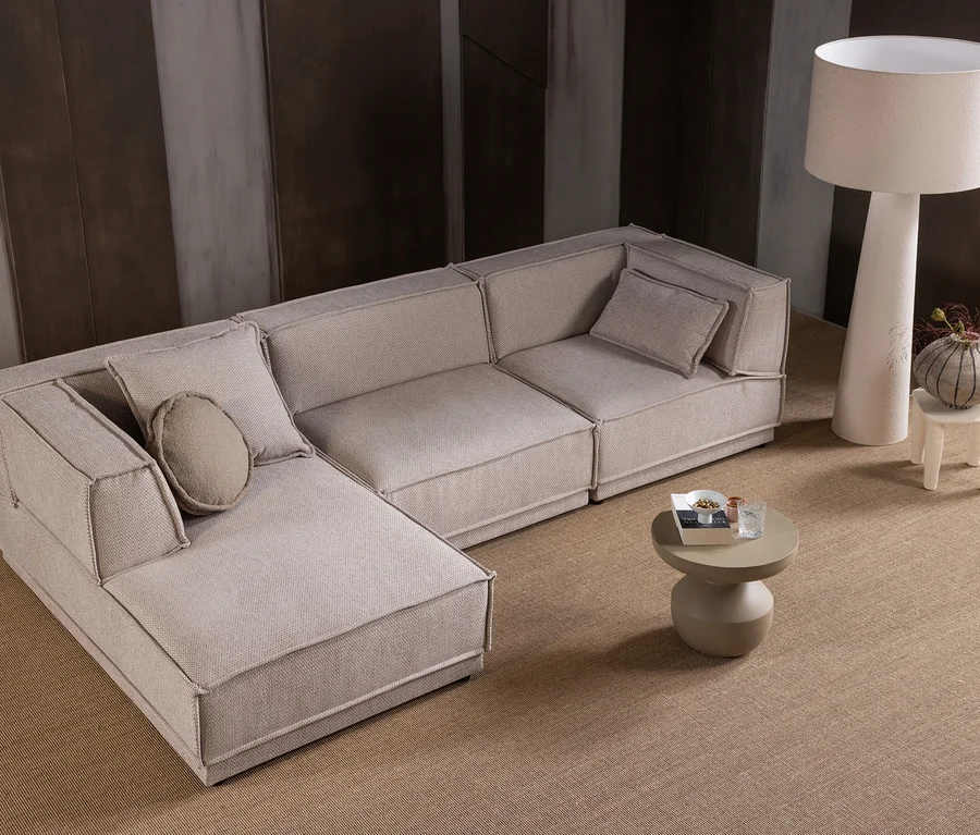 Ein L-förmiges Modulsofa aus hellbeigem Stoff mit sichtbaren Nähten steht auf einem strukturierten braunen Teppich. Davor befindet sich ein kleiner runder Couchtisch mit einer Zeitschrift und einem Glas. Rechts davon steht eine hohe, cremefarbene Stehlampe neben einem kleinen weißen Hocker, auf dem eine gestreifte Vase mit Pflanzen steht. Im Hintergrund sind gestreifte Wände zu sehen.